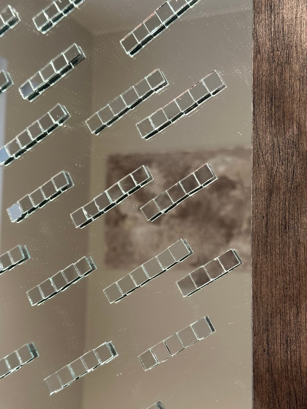Close-up of a mirror adorned with raised mirror mosaics, reflecting a textured beige painting by Anne Joris